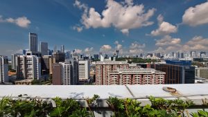 Panoramic city skyline viewed from a rooftop, with high-rise buildings, scattered clouds, and plants in the foreground.